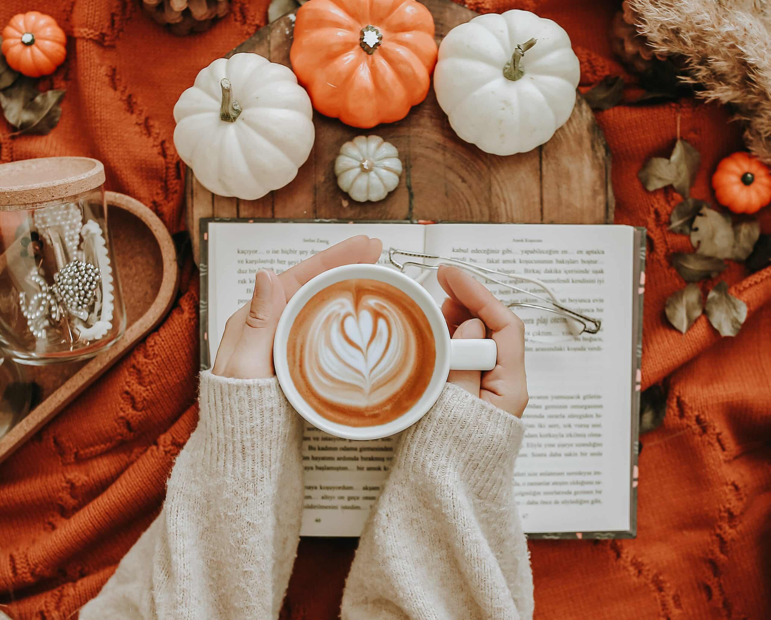 Picture of a woman's hands, sitting on top of an open book, holding a cup of cappuccino. On the table, there are orange and white pumpkins, an orange piece of fabric and other orange colored fall decor items.